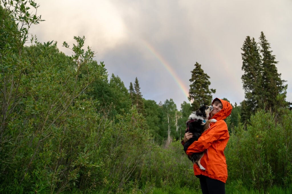 Kristen Bor wearing a red Arc'teryx Beta SL Rain Jacket holding her dog standing underneath a rainbow